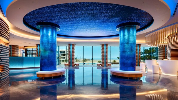 Area piscina interna moderna con soffitto a stelle, ampie colonne e vista sulla spiaggia.