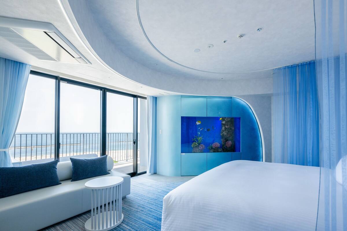 Camera d'albergo moderna con un letto grande, un'area salotto con vista sul mare e una parete azzurra a tema acquario.