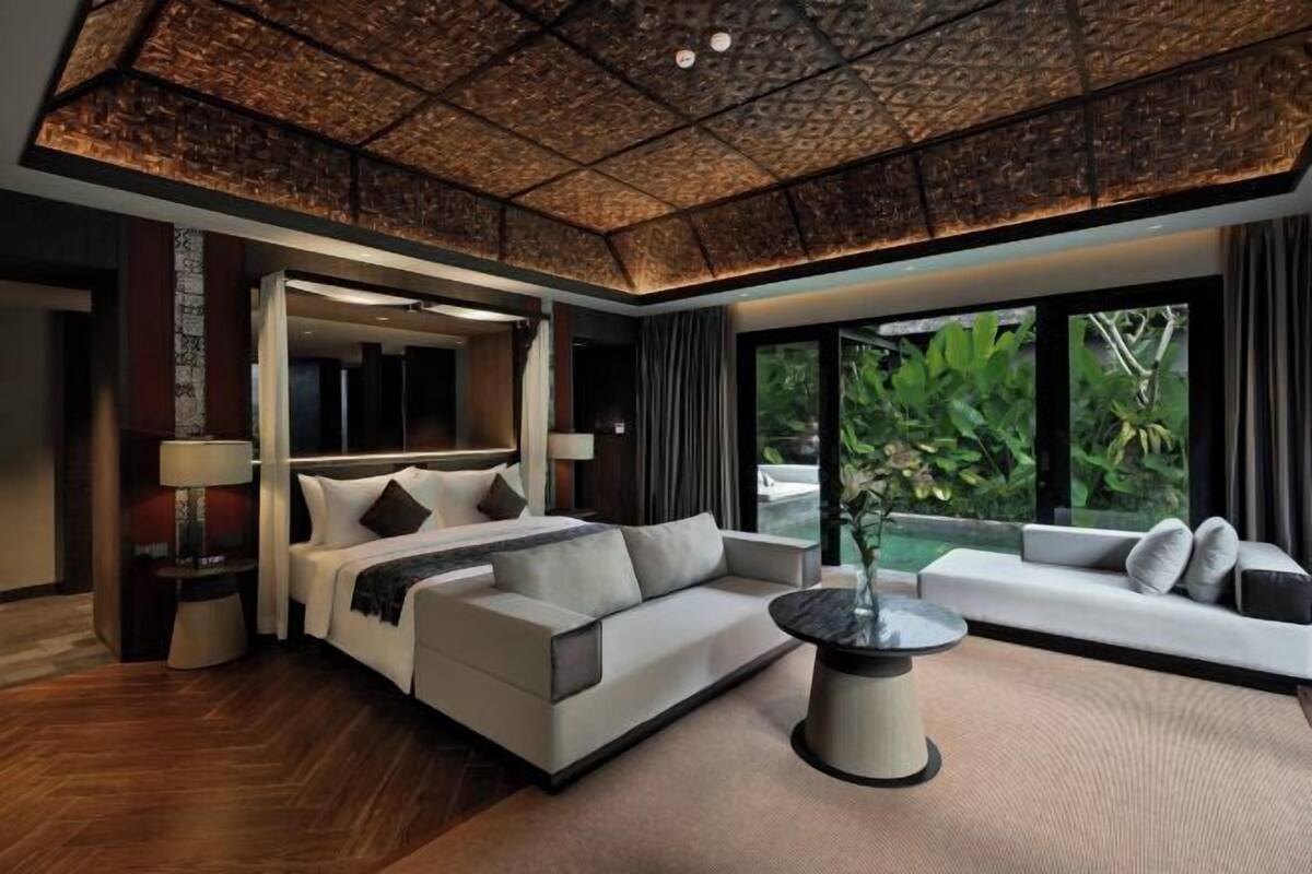 A modern hotel room with a large bed, a sofa, a coffee table, and a view of greenery outside.