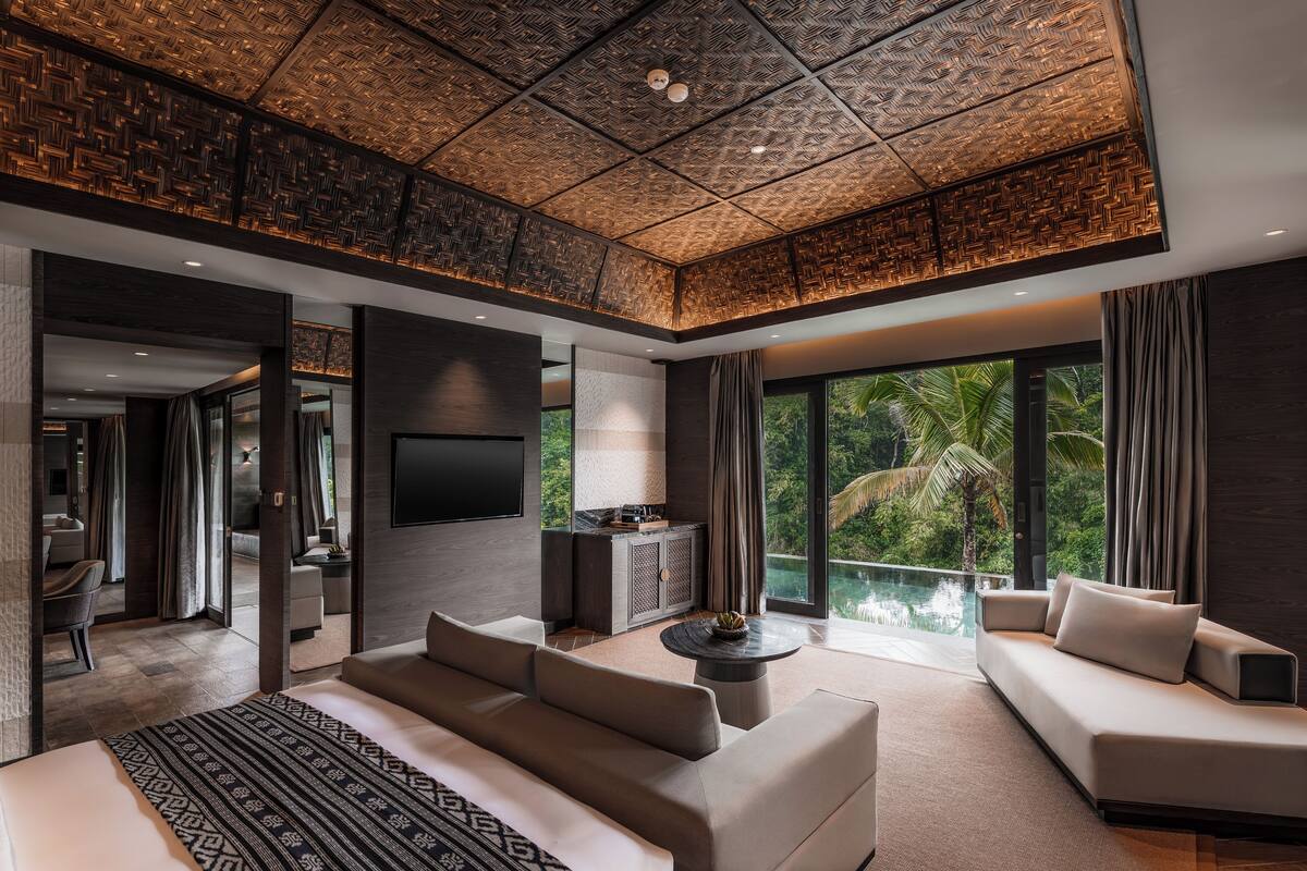 A modern living room with a large sofa, a coffee table, and a TV. There is a view of a pool and palm trees outside.