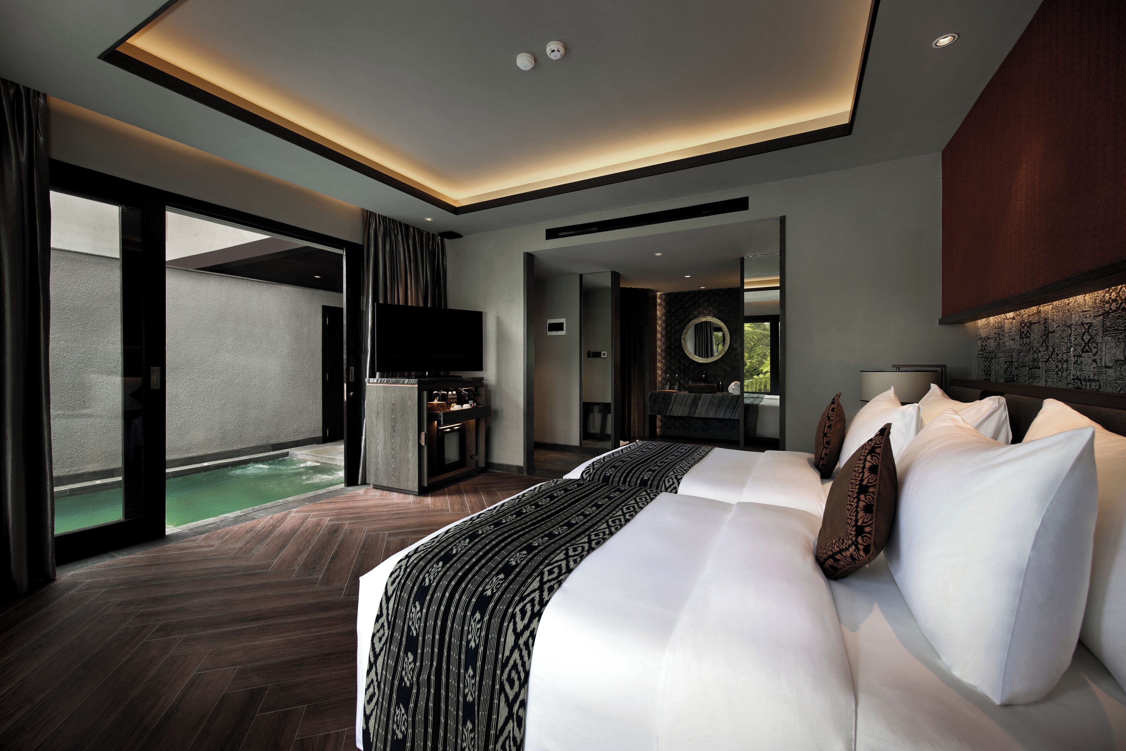 A modern hotel room with a large bed, a TV, a small kitchen area, and a view of a pool.