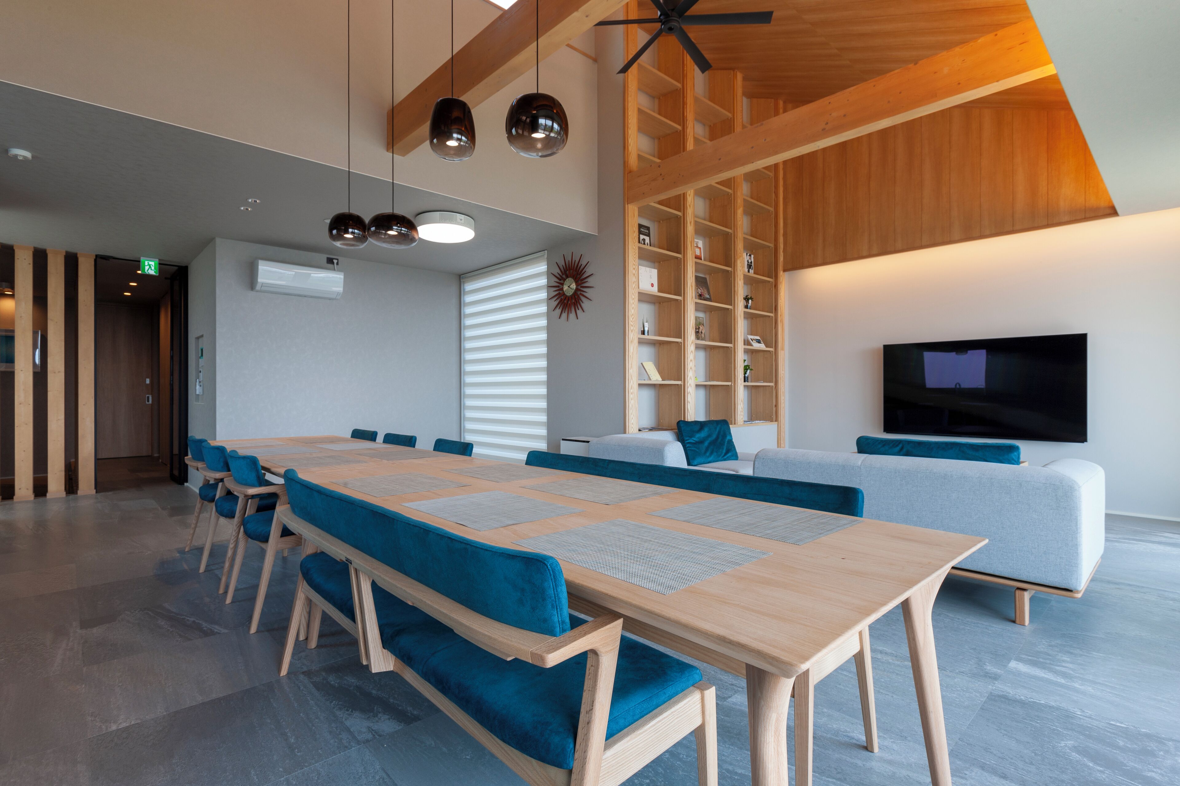 A modern interior with a long wooden table, blue upholstered chairs, a grey sofa, and a flat-screen TV mounted on the wall.