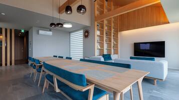 A modern interior with a long wooden table, blue upholstered chairs, a grey sofa, and a flat-screen TV mounted on the wall.