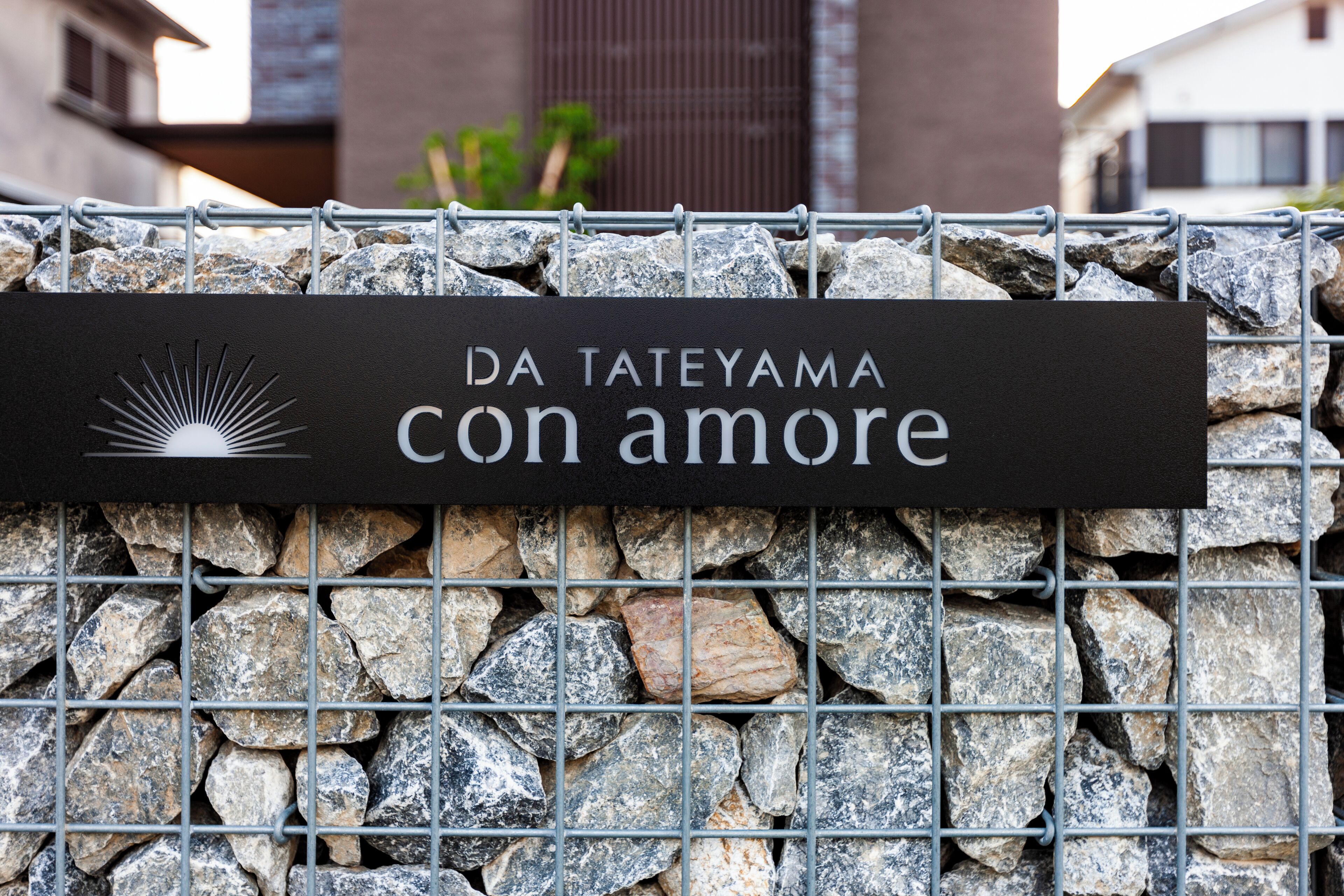 Un cartel con el texto "DA TATEYAMA con amore" en una valla metálica, con un muro de piedra al fondo.