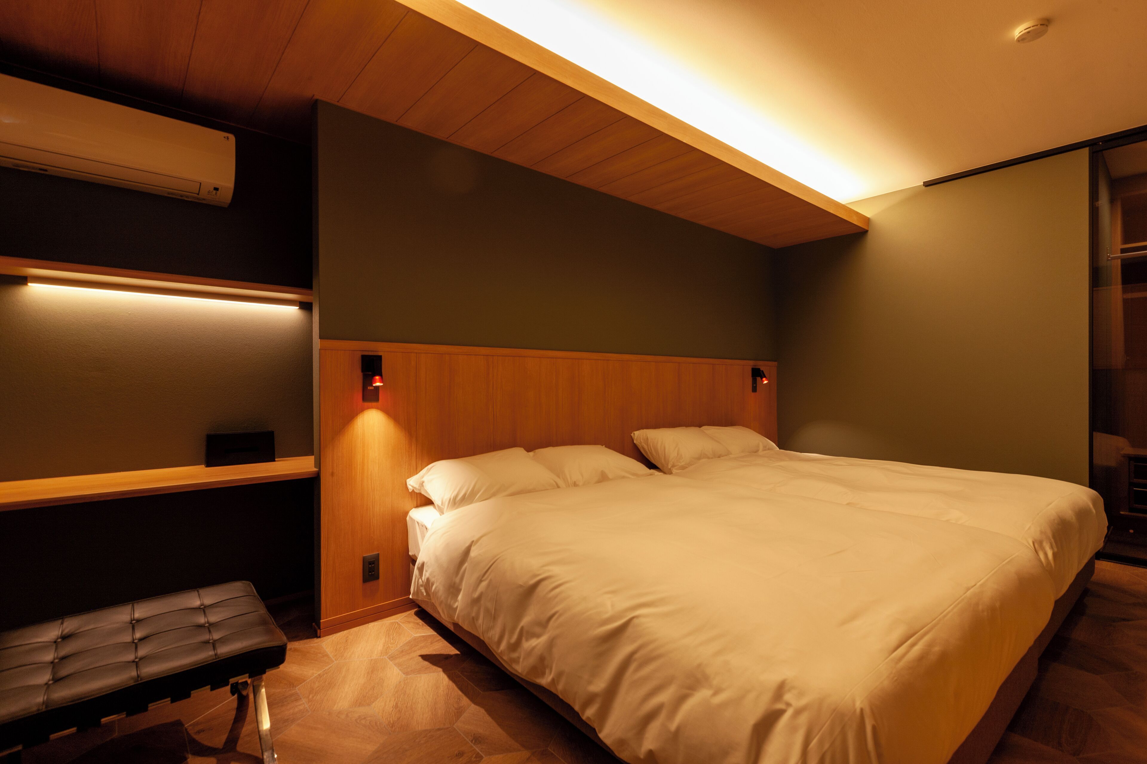A modern bedroom with a large bed, a bench, and a wall-mounted air conditioner.