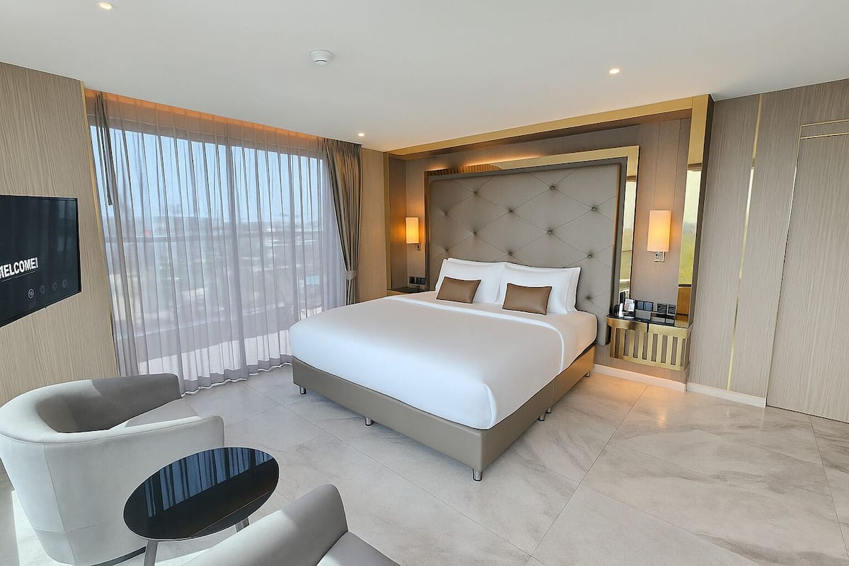 A modern hotel room with a large bed, a TV, a seating area with a table, and a view of the city through sheer curtains.
