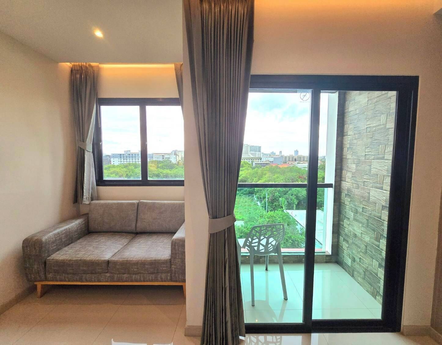 A room with a grey sofa, a chair, and a balcony with a view of buildings and greenery.