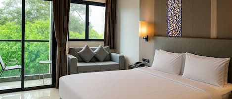 A hotel room with a large bed, a sofa, a small table, and a view of greenery through the windows.