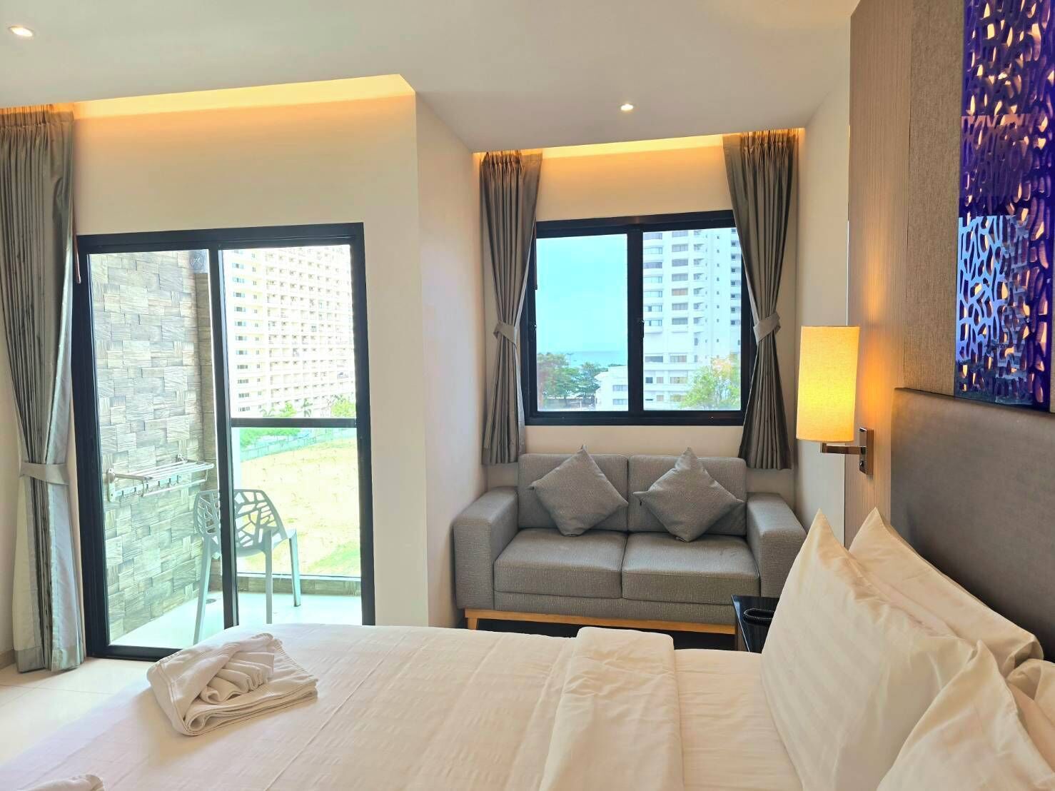 A hotel room with a bed, a sofa, a window with curtains, and a view of buildings.