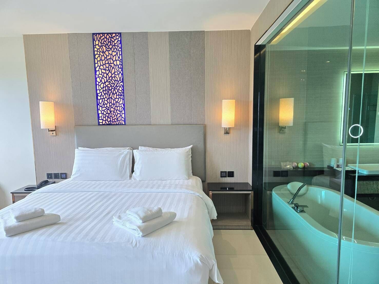 Ein modernes Hotelzimmer mit einem großen Bett, zwei Nachttischen, einer dekorativen Wandverkleidung und einem verglasten Badezimmer mit Badewanne und Spiegel.