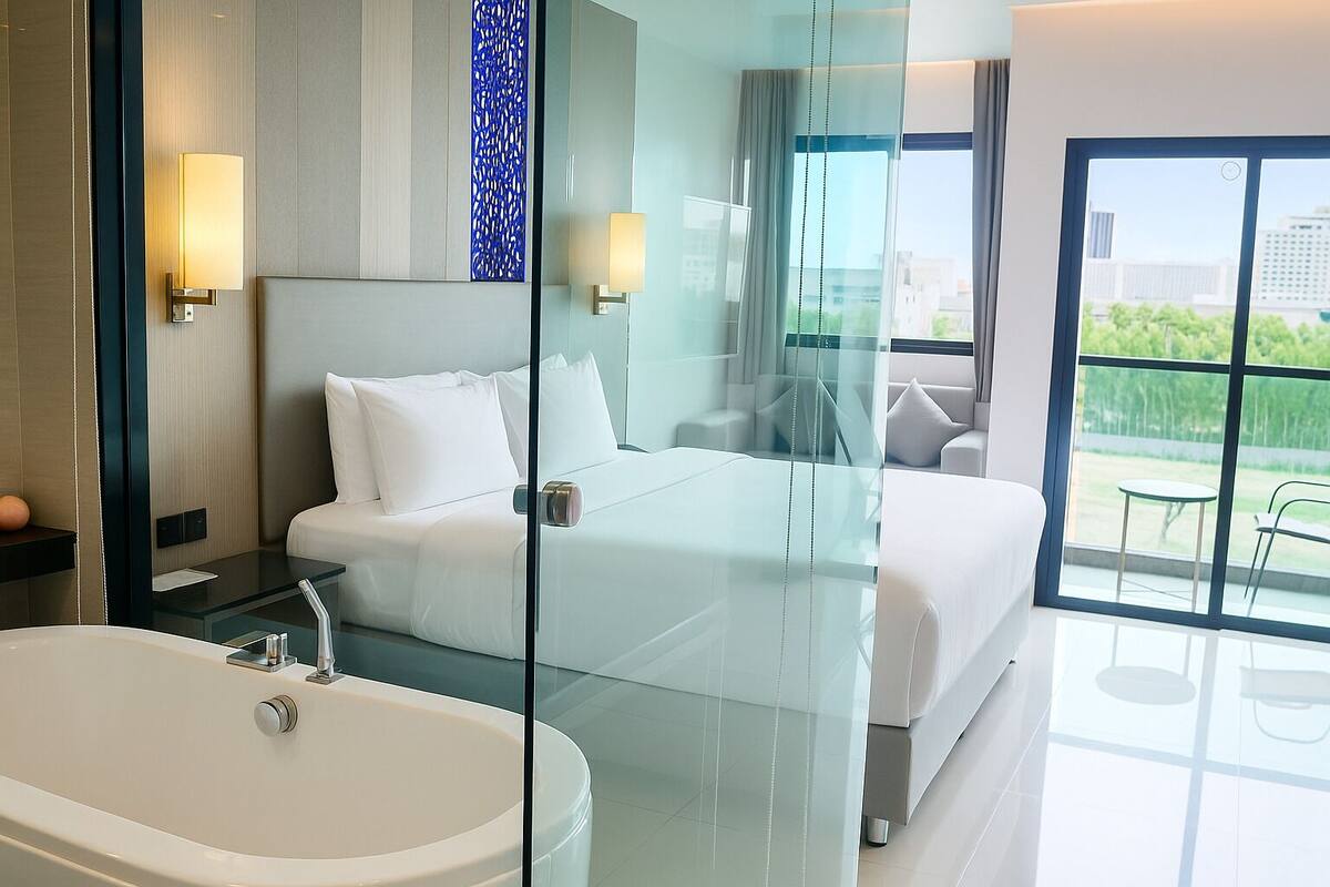 A modern hotel room with a glass-enclosed bed, a large window with a view of the city, a separate bathroom with a bathtub, and a small balcony with chairs.