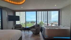 Una habitación de hotel moderna con una cama grande, un área de descanso con sillas, una mesita y una bañera amplia, además de un televisor montado en la pared.