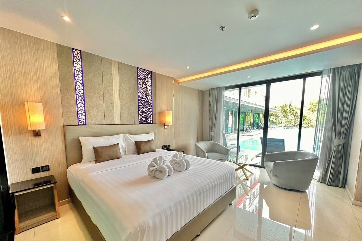 A modern hotel room with a large bed, two bedside lamps, a glass table, a chair, and a view of a swimming pool.