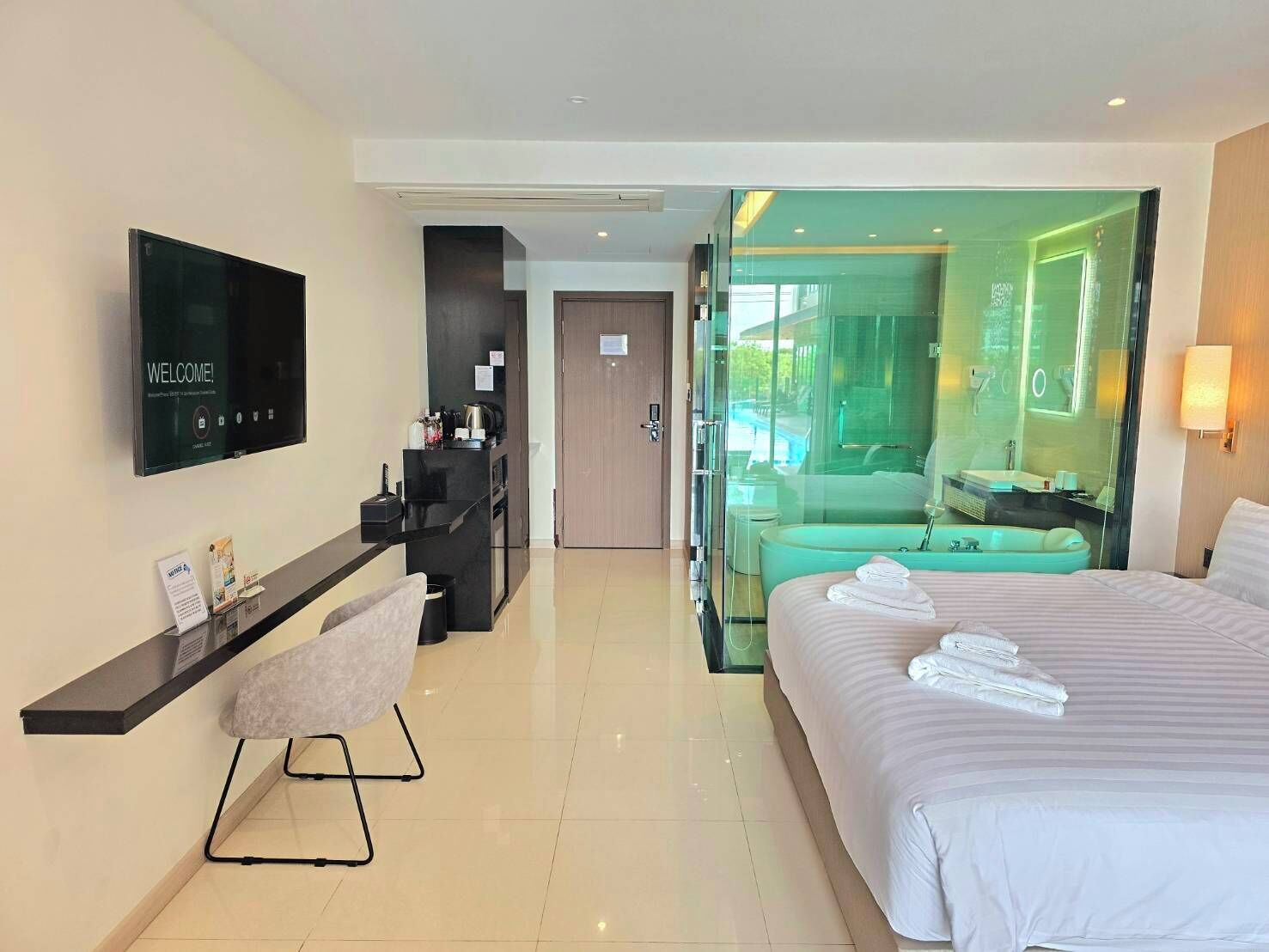 A modern hotel room with a bed, a desk, a chair, a television, and a glass-enclosed bathroom.