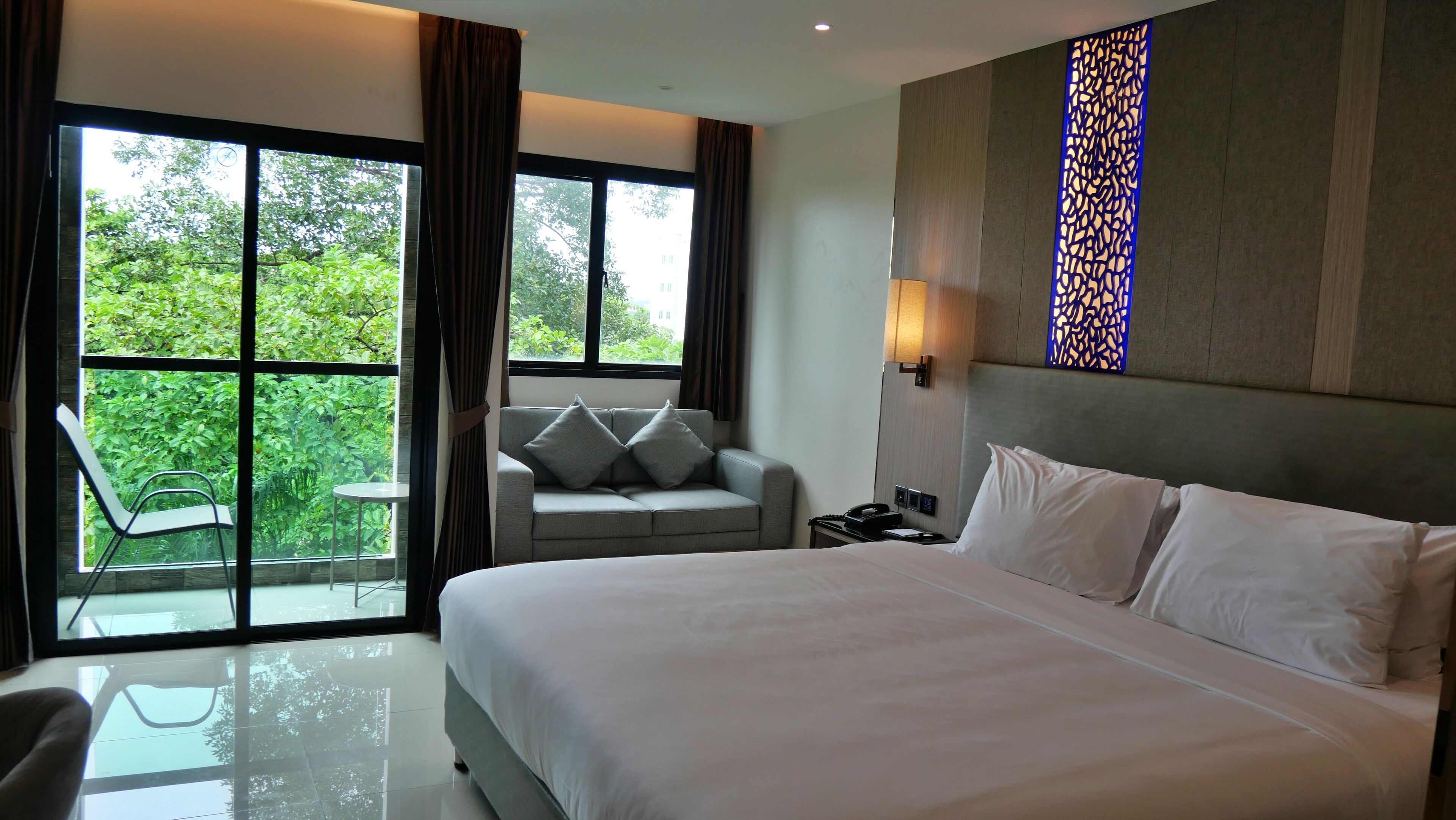 A hotel room with a large bed, a sofa, a chair, and a view of greenery through the windows.