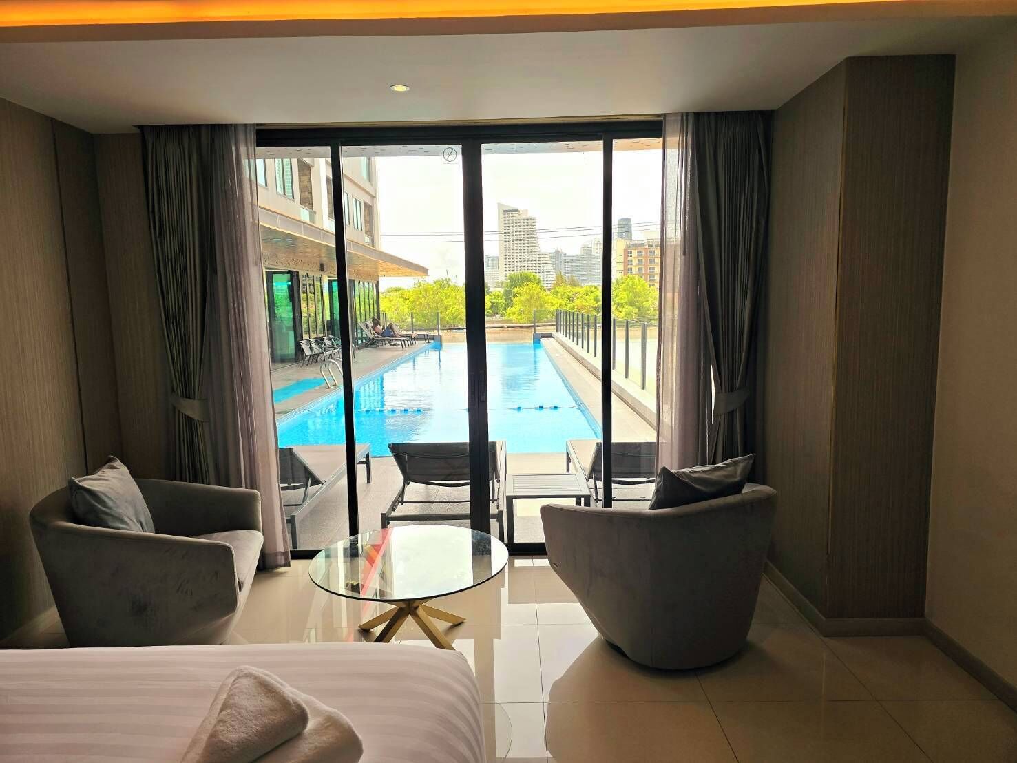 A modern hotel room with a large bed, two armchairs, a glass coffee table, and a view of an outdoor swimming pool and cityscape.