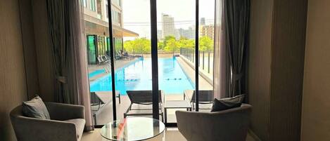 A modern hotel room with a large bed, two armchairs, a glass coffee table, and a view of an outdoor swimming pool and cityscape.