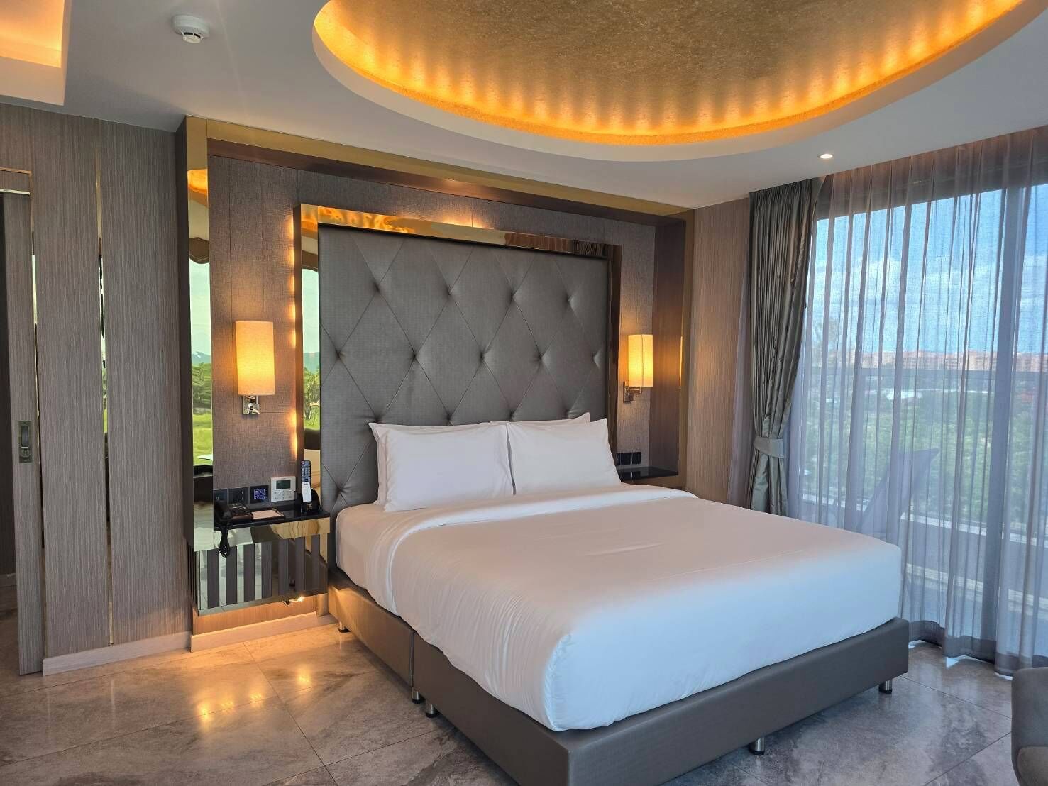A modern hotel room with a large bed, a tufted headboard, bedside lamps, and a view of the outdoors through large windows.