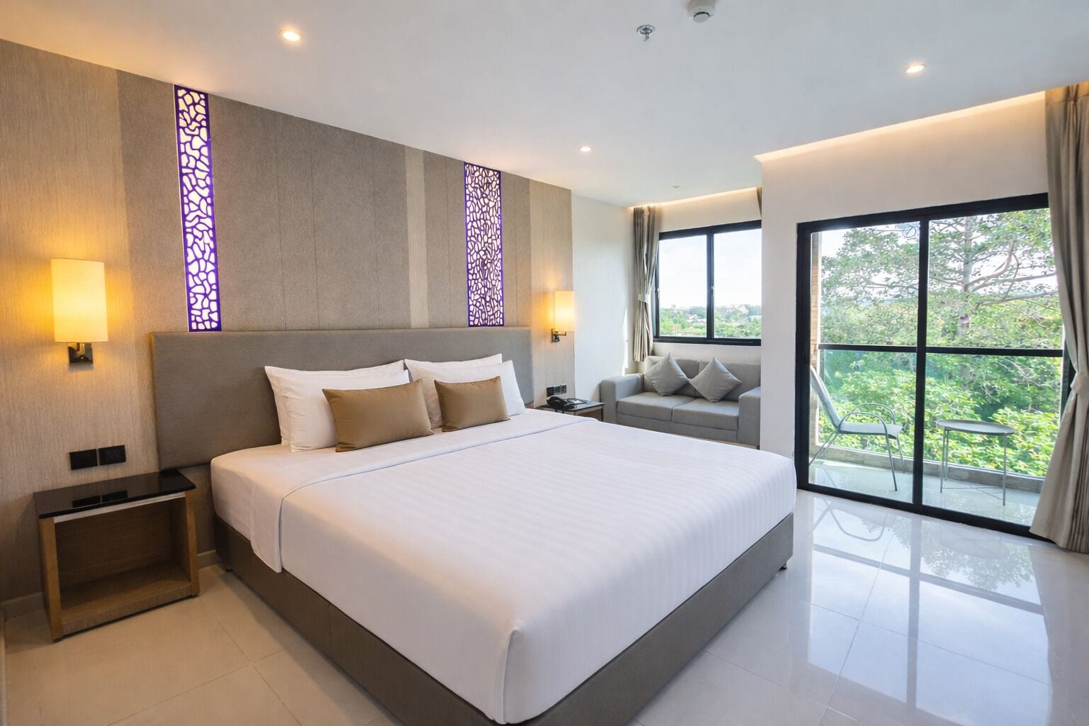 A modern hotel room with a large bed, bedside lamps, a sofa, and a view of greenery through large windows.