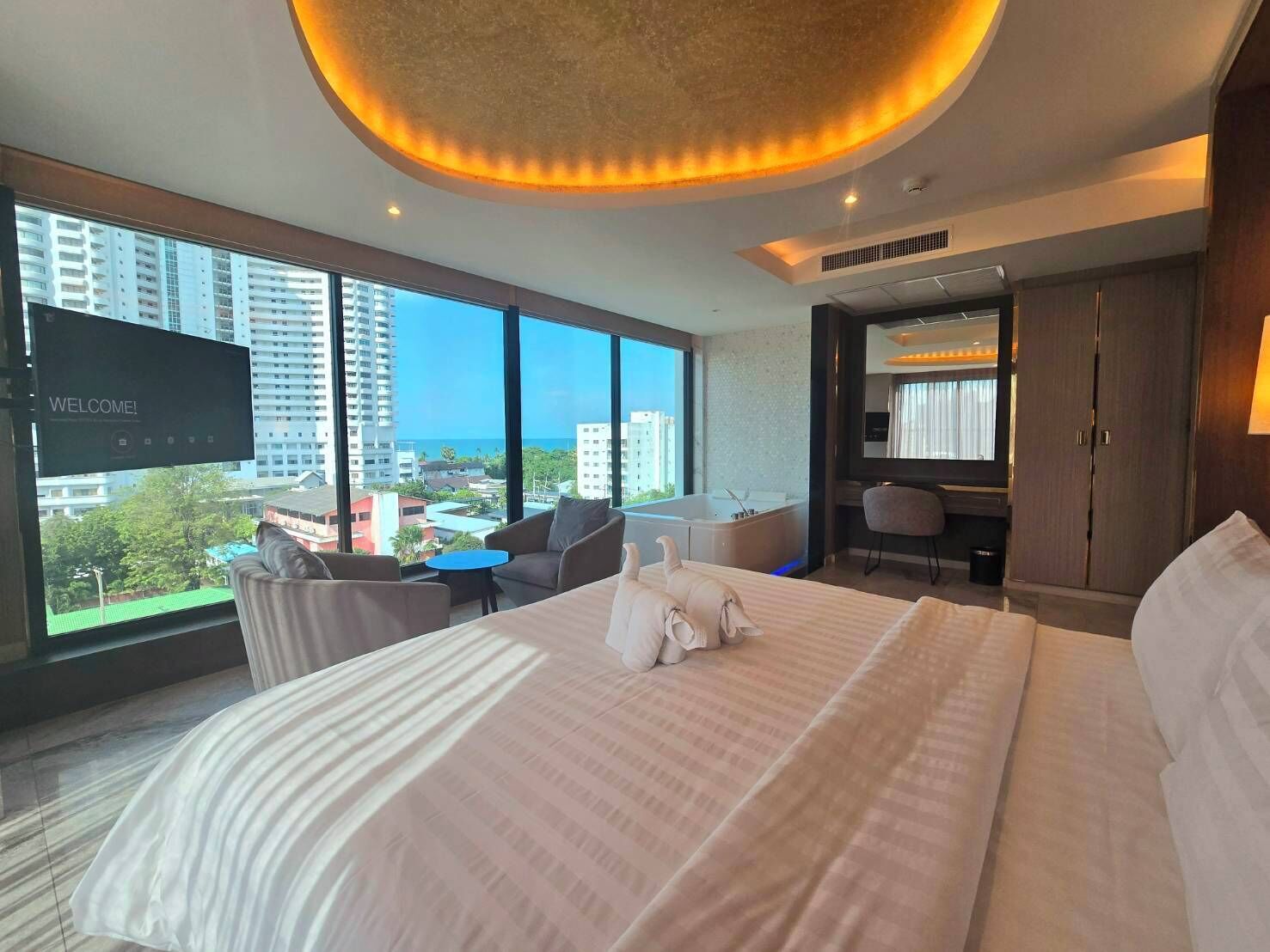A modern hotel room with a large bed, a desk, a chair, and a TV displaying "WELCOME!" The room has large windows offering a view of the city and buildings.