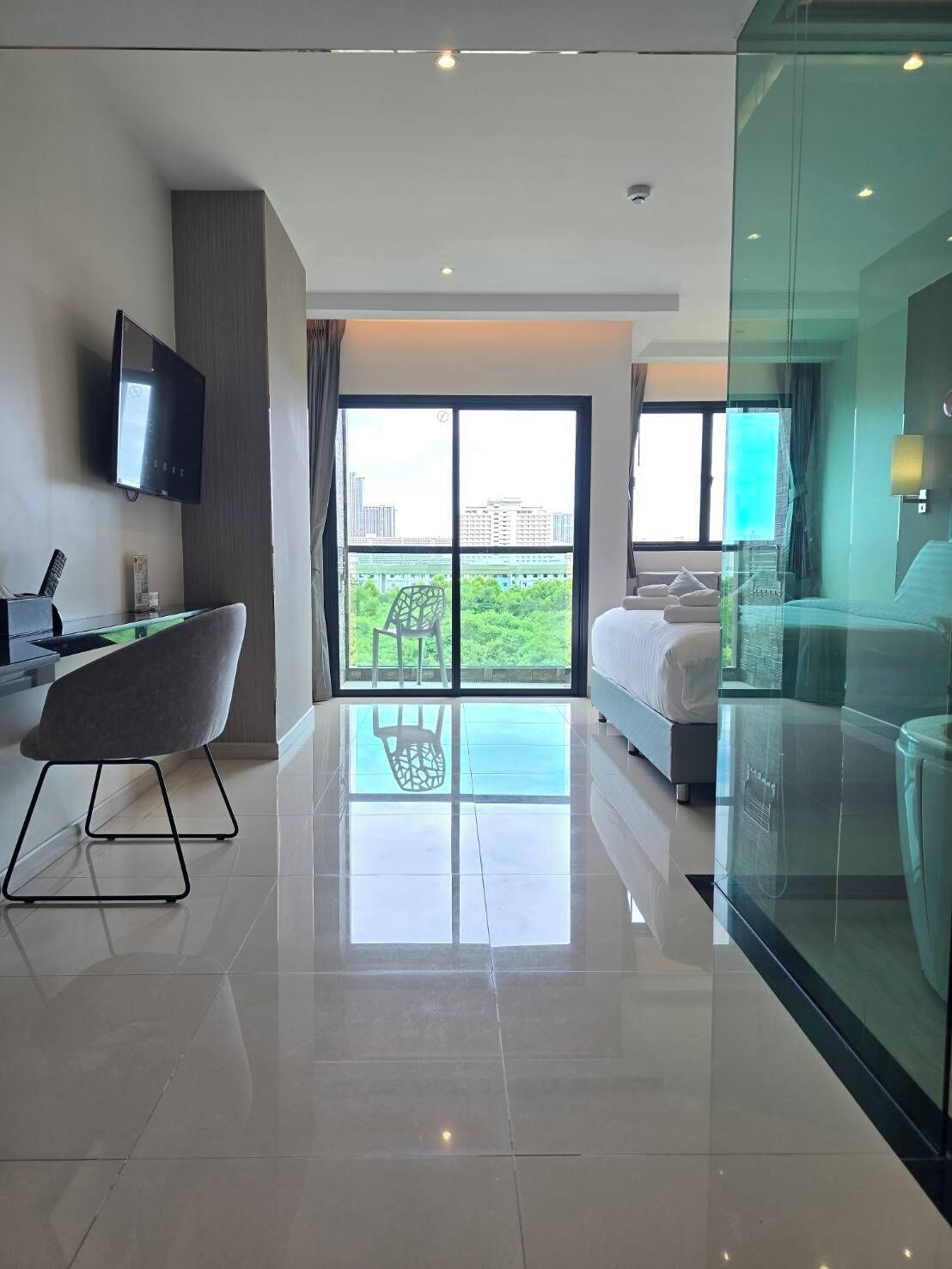 A modern hotel room with a large sliding glass door to an outdoor balcony, a flat-screen TV mounted on the wall, a desk with a chair, and a bed with a nightstand.