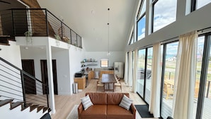 A spacious living area with a high ceiling, a wooden staircase, a dining area with a table and chairs, and large windows offering a view of the outdoors. - Bryce, Zion National and State Park Retreat (Hatch)