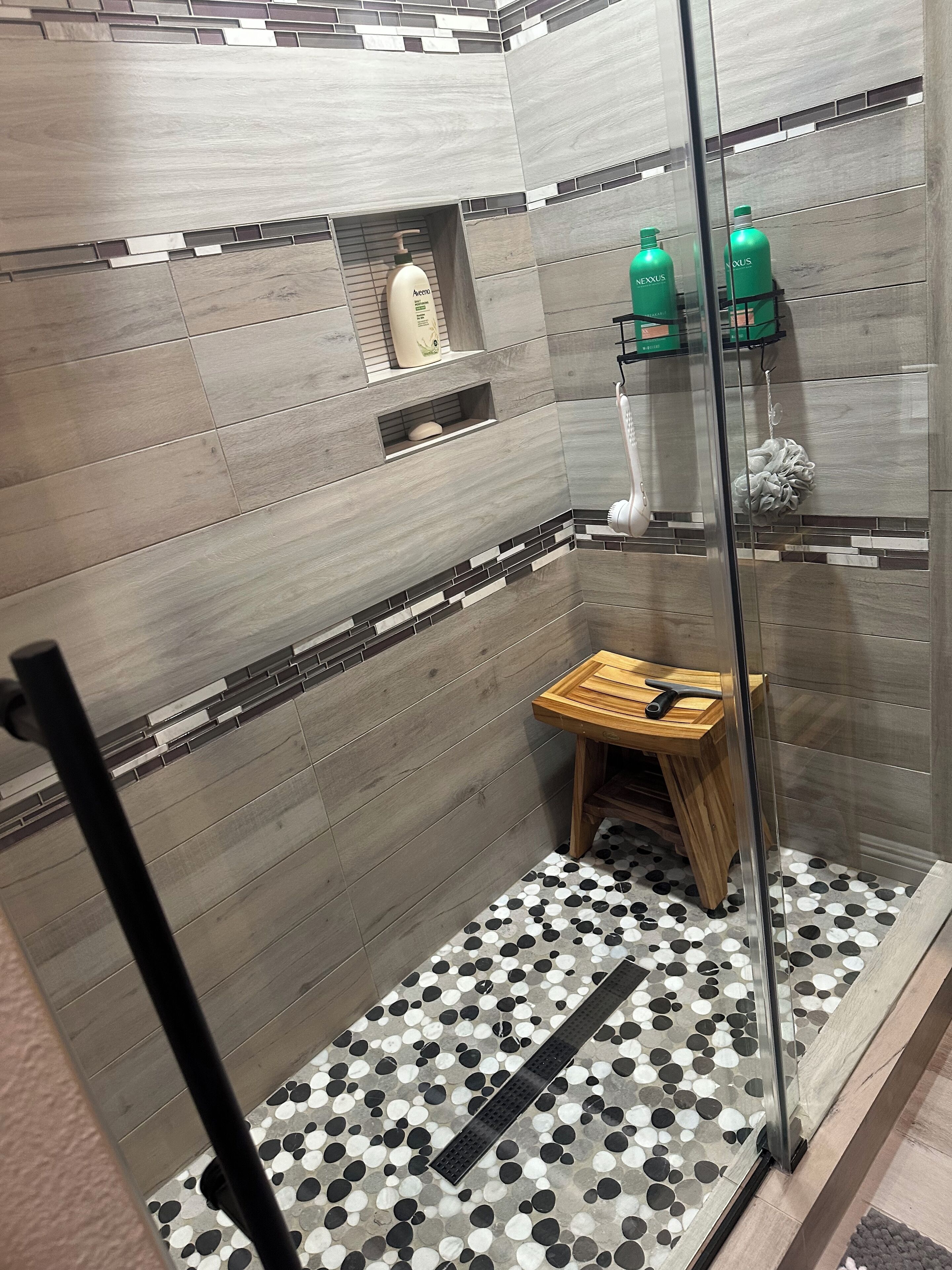 A modern shower area with grey tiled walls, a wooden bench, and a black and white patterned floor.