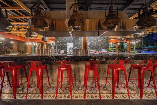 Una zona de bar con taburetes rojos, mostrador de bar de acero inoxidable e iluminación industrial suspendida.