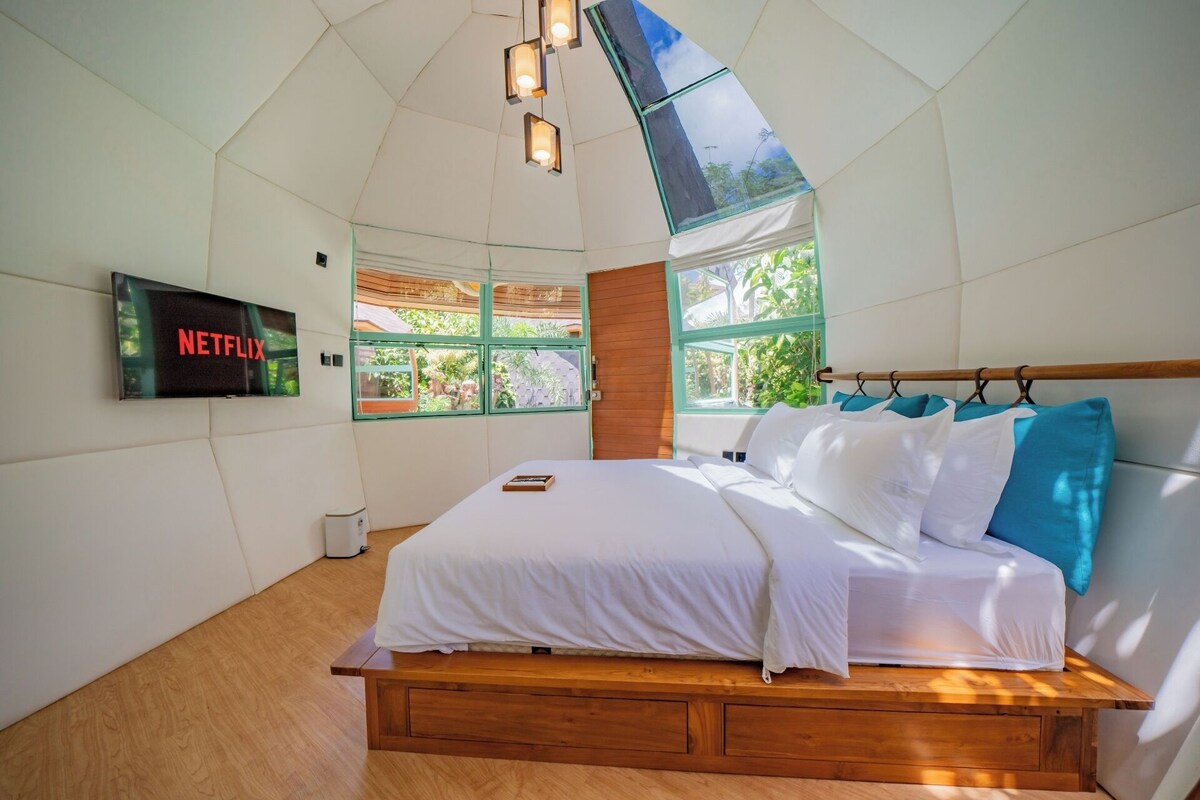 A modern bedroom with a wooden bed, a TV mounted on the wall, and large windows offering a view of greenery.
