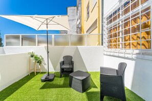Un patio con césped artificial, mobiliario exterior, un gran paraguas y un edificio con una ventana al fondo.