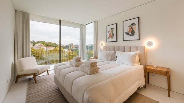 A modern bedroom with a large bed, a chair, a bedside table, and a view of the city.