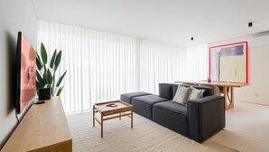 A modern living room with a grey sofa, a wooden coffee table, a TV on a stand, and a large window with blinds.