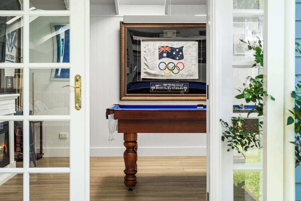 A room with a wooden table displaying a framed Olympic flag and memorabilia, a bookshelf, and a view through an open door. - Spa & Chill Lodge Budgewoi 3BR (Budgewoi)