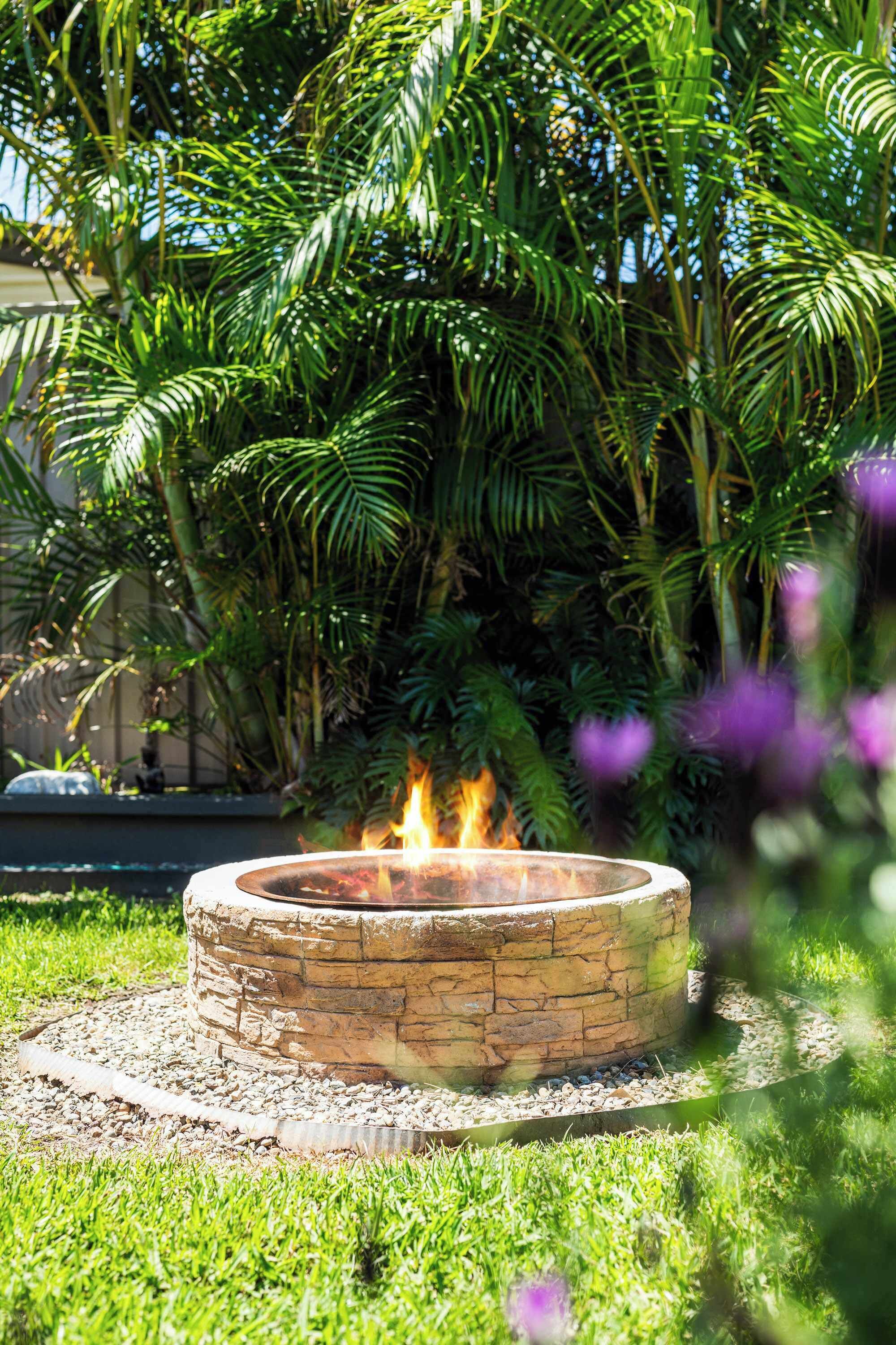 A circular stone fire pit surrounded by gravel, set in a grassy area with dense tropical foliage in the background.
