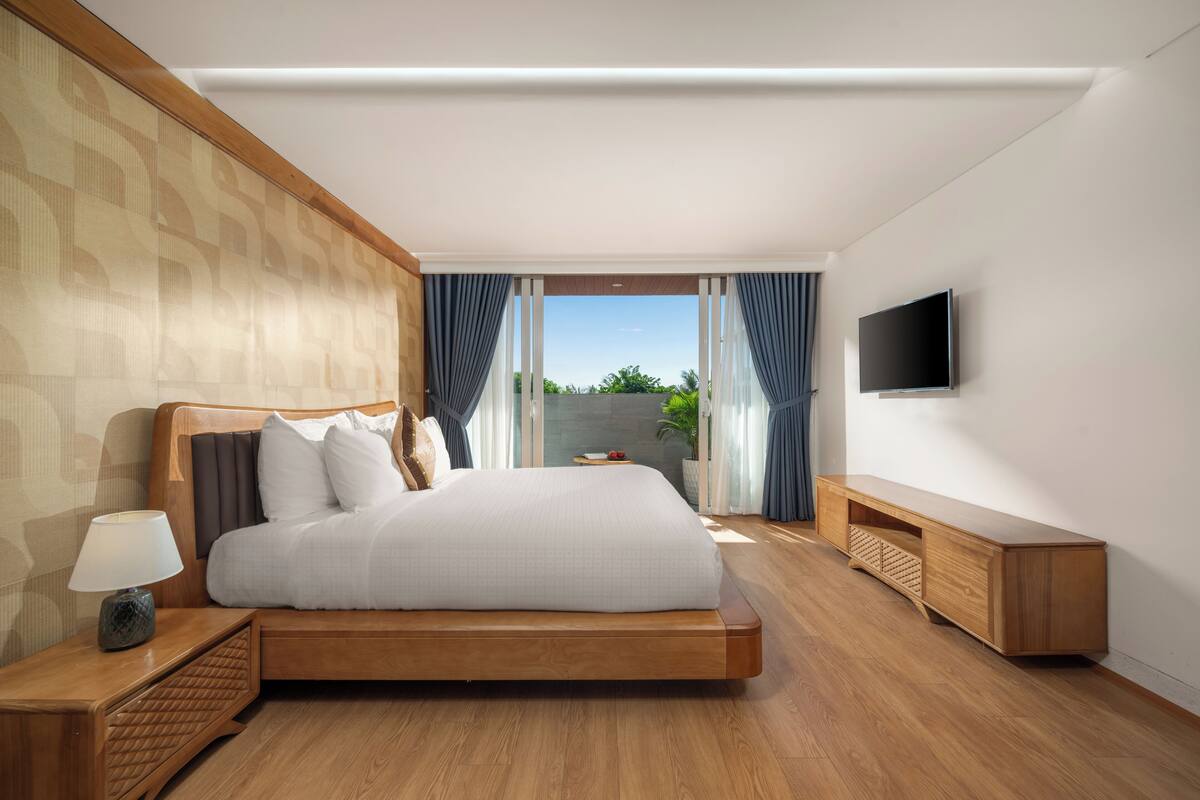 Une chambre moderne avec un grand lit, une tête de lit en bois, une table de chevet avec une lampe, un téléviseur fixé au mur et une vue sur l’extérieur grâce à une porte coulissante.