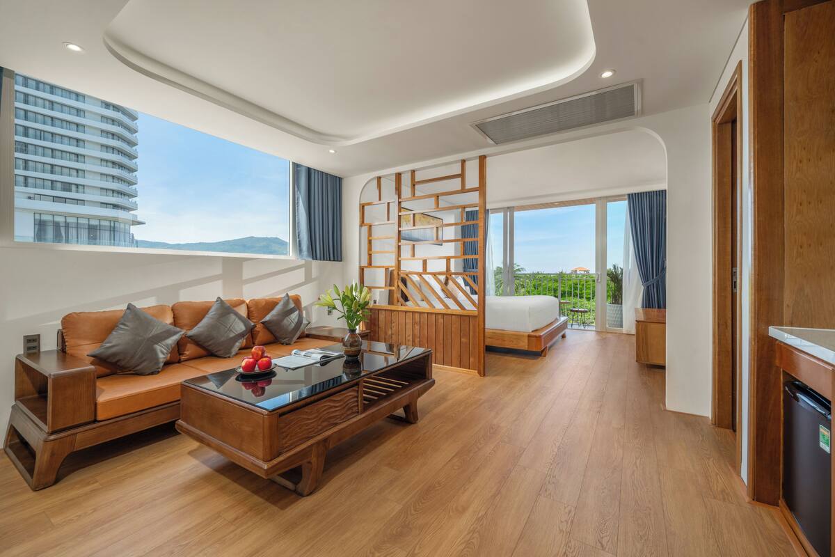 Une chambre d’hôtel moderne avec un canapé en bois, une table basse en verre, un bureau en bois et une grande fenêtre donnant sur un immeuble et de la verdure.