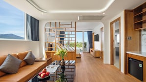 A modern hotel room with a wooden floor, a sofa with cushions, a coffee table with apples and a vase, a TV, and a kitchen area.