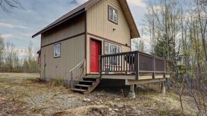 A two-story wooden cabin with a red door, a front porch, and a small staircase leading to the entrance. - The Northern Lights Oasis (Wasilla)