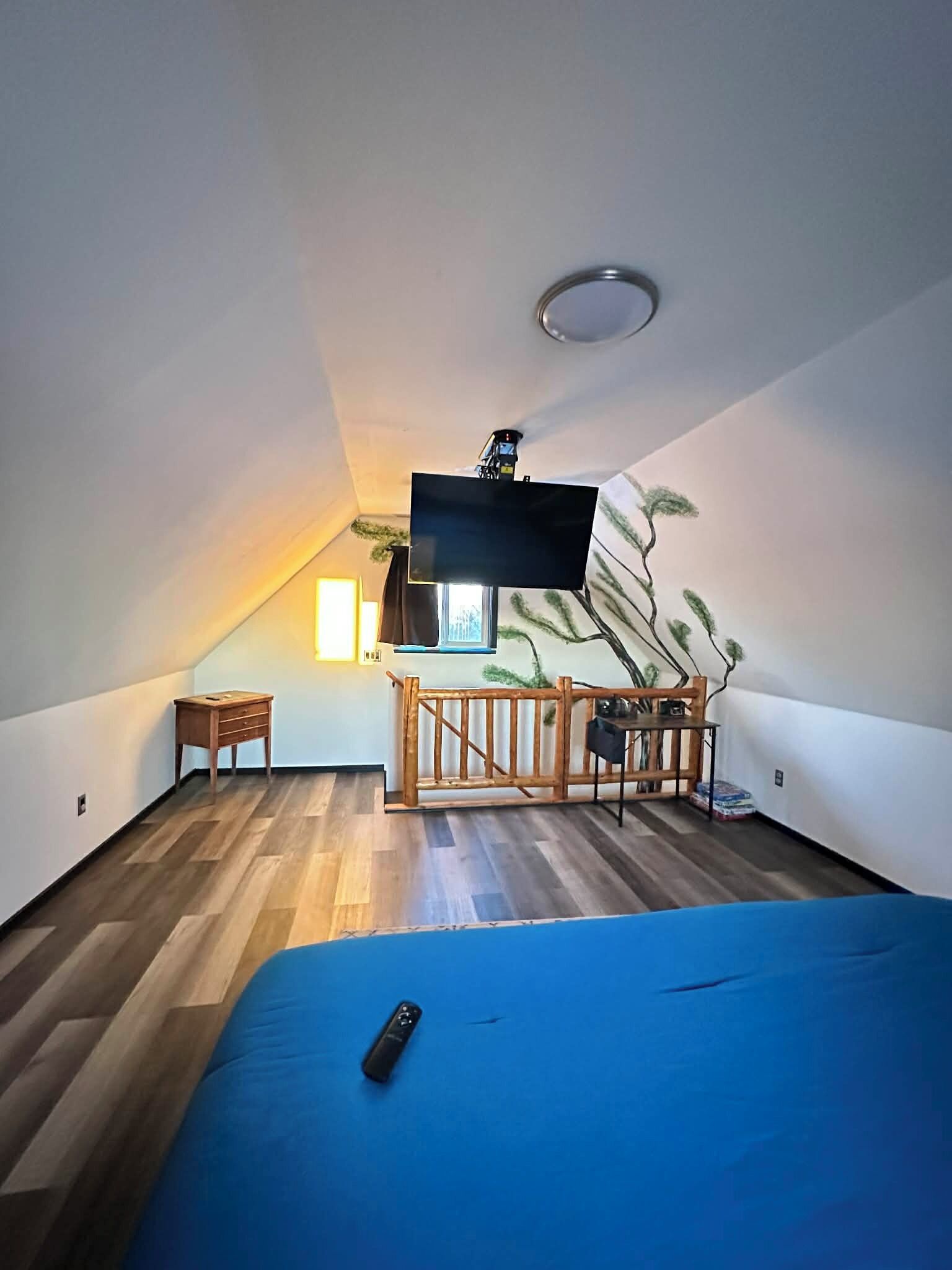 A room with a sloped ceiling, a bed, a TV mounted on the wall, a small table, and a chair.