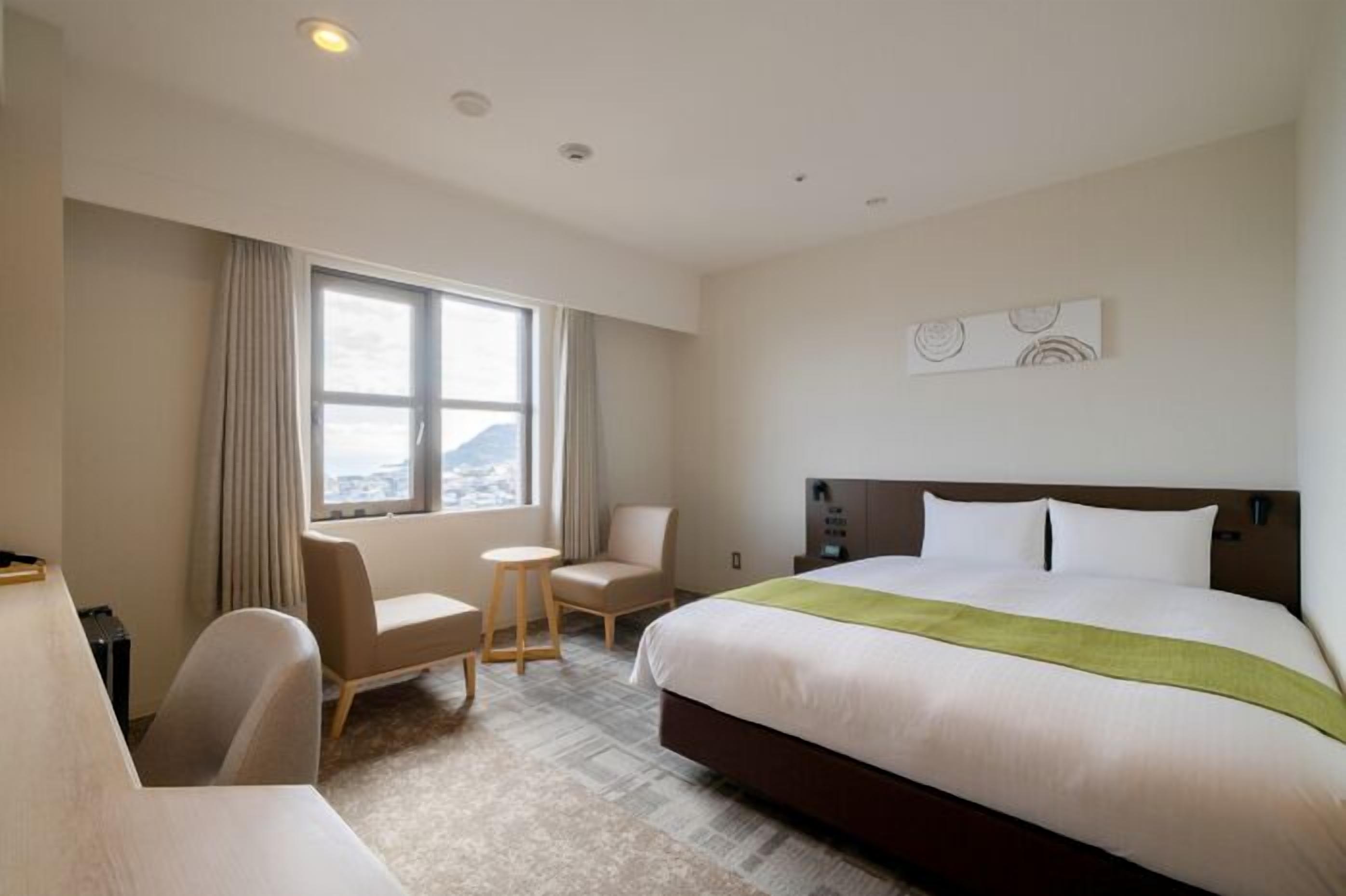 A hotel room with a bed, a desk, a chair, a window with curtains, and a city view.