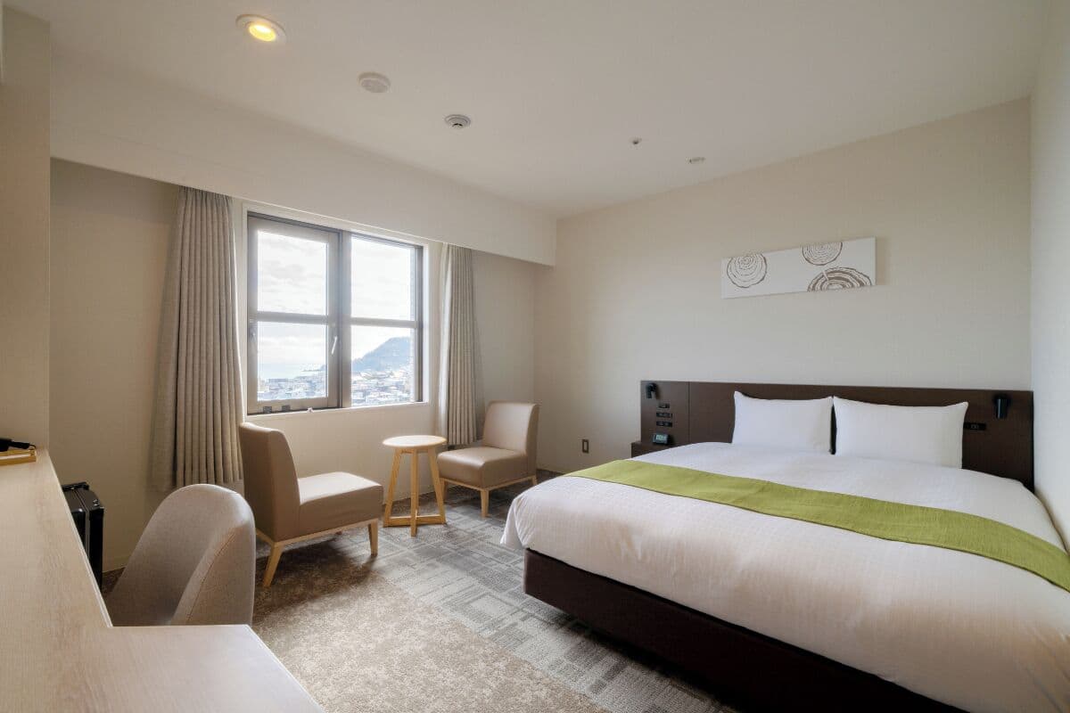A hotel room with a large bed, a desk, a chair, a small table, and a window with curtains.