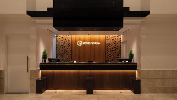 A hotel reception area with a wooden reception desk, a logo above it, and two potted plants on either side. - Hotel Enoe Hakodate (Hakodate)