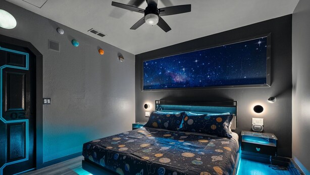 Ein Schlafzimmer mit Bett, Deckenventilator, Wand-TV mit Sternenhimmel und Nachttisch mit Lampe.
