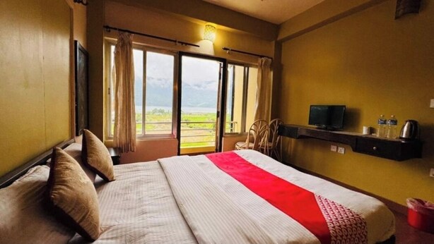A hotel room with a bed, a television, a shelf with items, and a view through a door. - Hotel August International (Pokhara)