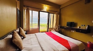 Habitación de hotel con cama, televisor, una estantería con objetos y vista por una puerta.