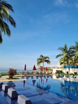 Zona de piscina con sillas de descanso y sombrillas, palmeras y vista al mar.