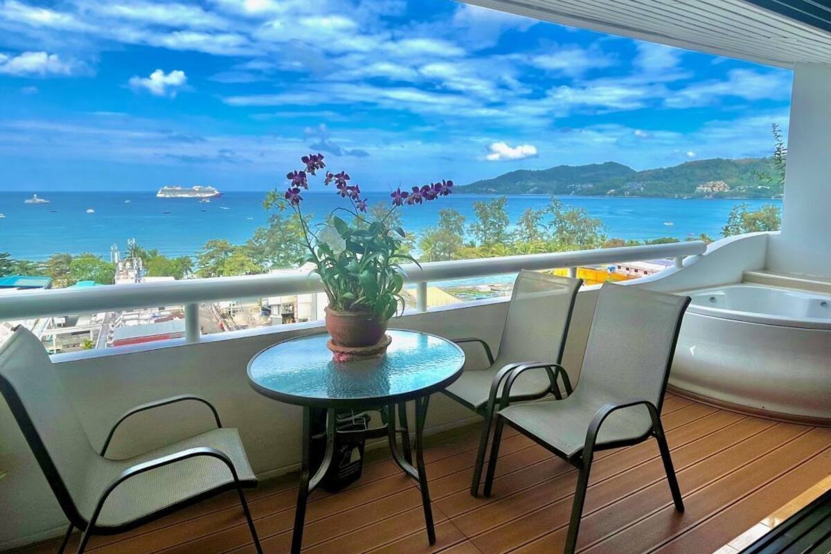 Ein Balkon mit rundem Tisch, zwei Stühlen, einem Blumentopf und Blick auf das Meer mit Booten und Hügeln.