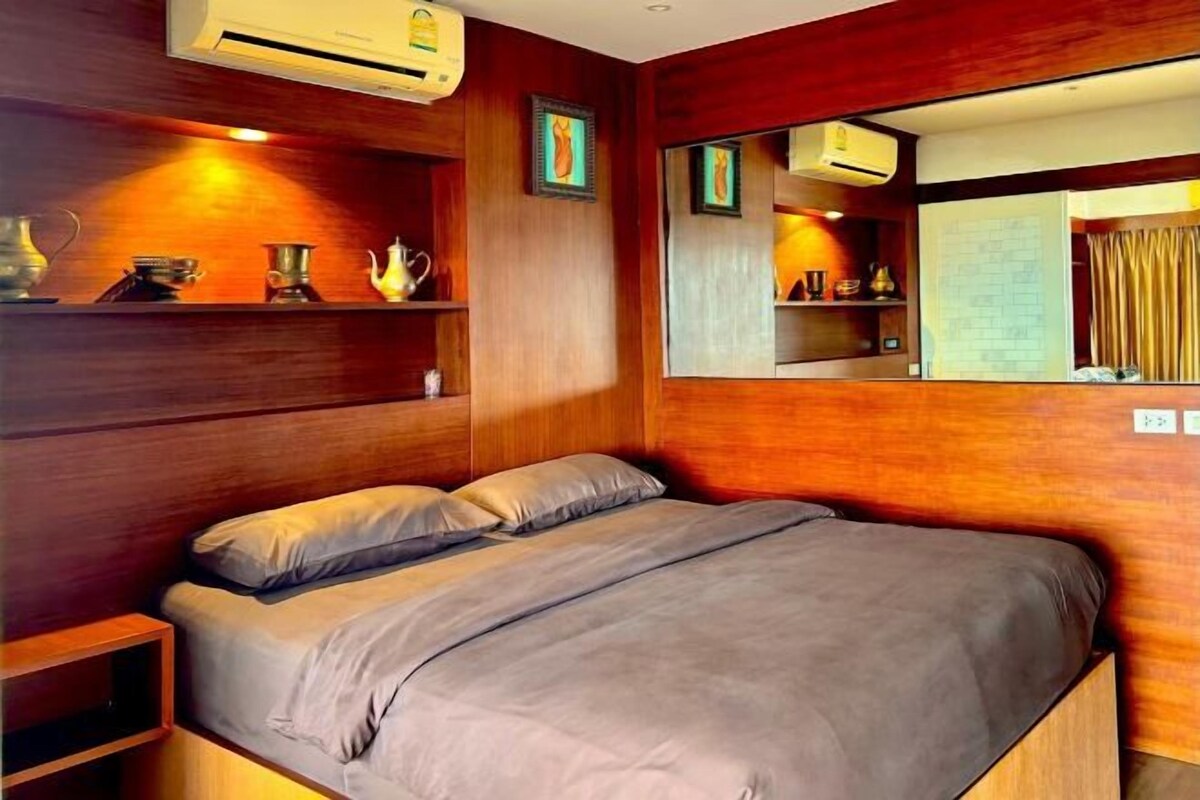 A bedroom with a wooden headboard, a bed with grey bedding, a bedside table, and a mirror on the wall.