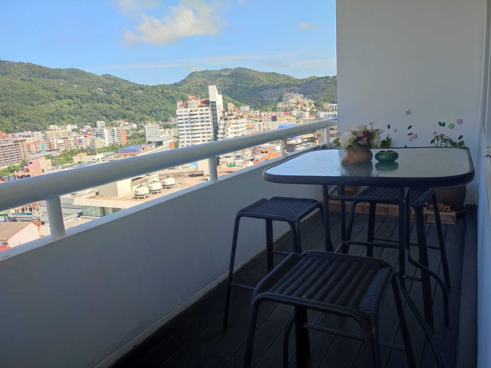 A balcony with a table and two chairs, overlooking a cityscape with buildings and hills.