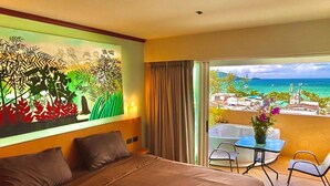 Habitación de hotel con cama, un cuadro, balcón con mesa y sillas, y vista al mar y edificios.